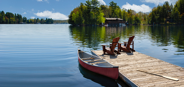Two Adirondack chairs on a wooden dock overlook the serene blue waters of a Muskoka lake in Ontario, Canada. Nearby, a red canoe is tied to the pier, alongside life jackets and a paddle. Space for text.
