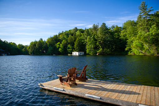 Two Adirondack chairs on a wooden dock on a summer morning in Muskoka provide a serene vista of the lake. Nestled among the trees across the water, cottages complete the picturesque scene.
