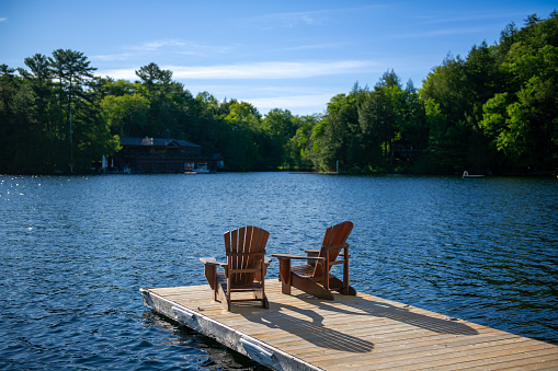 Soaking in the sunshine of a bright Muskoka morning, two Adirondack chairs peacefully rest on a wooden dock, surveying the tranquil waters of the lake.