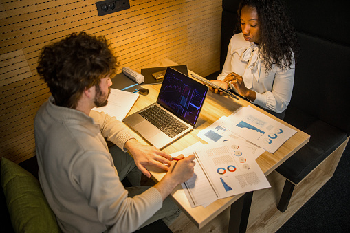 A Hispanic male and Black female, dressed in professional attire, scrutinize financial charts on a laptop and documents, indicating their concentrated collaboration in a modern office setting.