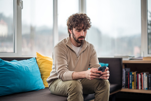 A Hispanic male investor dressed in casual attire intently reviews financial data on his smartphone, seated comfortably indoors with sunlight filtering through the windows.
