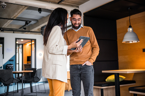 A smiling Hispanic man in a sweater shares a tablet with a professional female partner in a chic office setting, discussing investment opportunities.