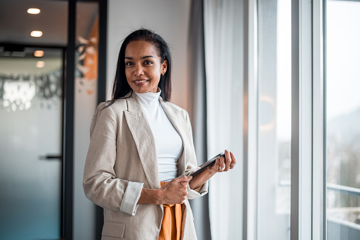 A mature Hispanic woman exudes professionalism in a modern office, smartly attired as she holds a digital tablet, symbolizing her role as a savvy investor.