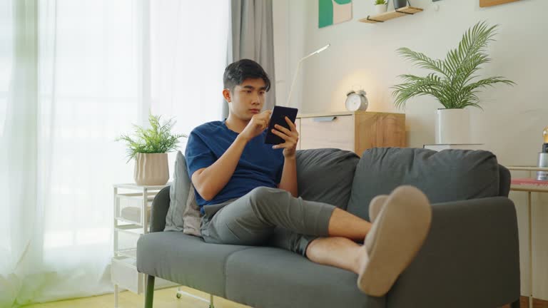 Asian man relaxing reading on E-book holding a tablet displaying text on sofa in living room at home ,modern lifestyle