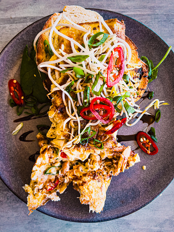 Vertical high angle closeup photo of an omelette with crab pieces, shallots, brown rice vinegar, bean sprouts and sliced red chilli on toasted sourdough bread, served on a dark grey ceramic plate in a cafe restaurant.