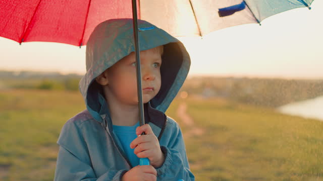 Upset kid with umbrella at rain on riverbank