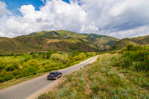 A car is cruising along an asphalt road in the countryside with mountains towering in the background, under a clear blue sky with fluffy clouds