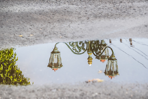 Reflection of typical street lamp from Yogyakarta on water surface on asphalt road after rain. Classic Javanese green framed glass lamp against bright blue sky background. Empty blank copy text space.