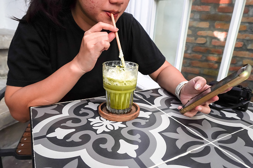 A woman is checking social media while drinking a sweet matcha drink with cream in a cafe. Someone who is addicted to gadgets using smartphone or mobile cell phone. Concept for unhealthy lifestyle.