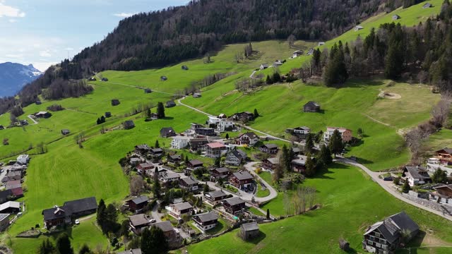 Small Swiss Village on green mountain during sunny day in spring. Picturesque landscape in Switzerland. Aerial birds eye forward shot.