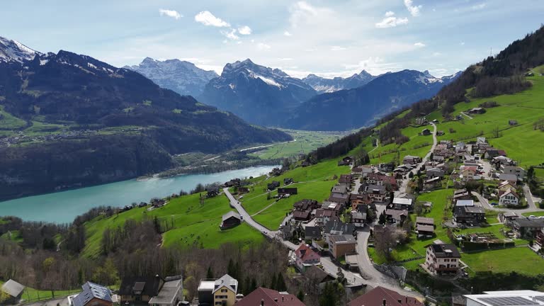 Aerial establishing shot of  idyllic swiss village named Amden during sunny day. Walensee Lake in the valley with quaint mountain range in background. Snowy summit on mountaintop. Wide shot.