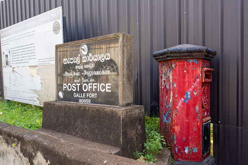 Galle, Sri Lanka - January 29, 2024: A vibrant red postbox stands on the streets of the historic Galle Fort, with a signboard written in local language and Post Office, Galle Fort next to it.