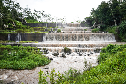 Grojogan Watu Purbo, Yogyakarta. Large multi-level dam on a river whose water is murky brownish due to mud during heavy rain surrounded by trees and meadows during overcast. Artificial Waterfall.