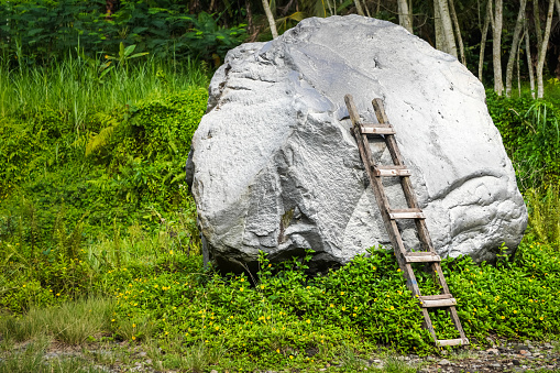 An old and rotten wooden ladder was propped up against a boulder. Wood ladder on solid stone wall backgrounds. Empty blank copy text space.