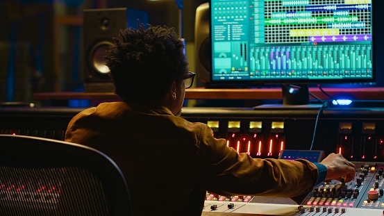 Sound designer uses mixing console and managing audio tracks, twisting knobs on control desk for adjusting tunes and volume. Producer creates music in professional studio, technical gear. Camera A.