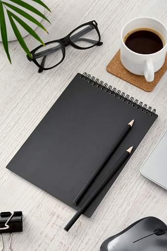 A black notebook sits on a desk with a pair of black pencils and a cup of coffee. The scene suggests a work environment where someone is likely to write or take notes