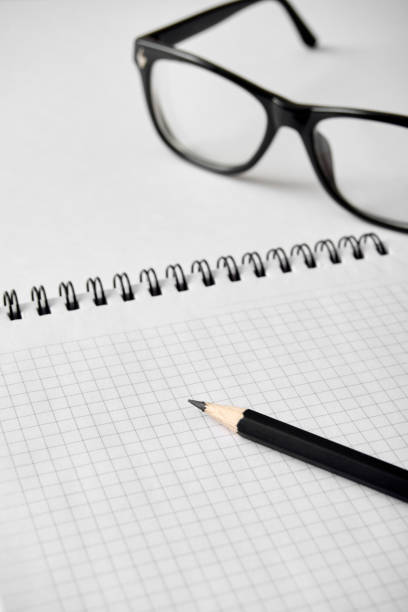 A close-up view of a pair of black eyeglasses lying beside a sharpened black pencil on an open notebook with graph-lined paper. The setup suggests a moment of pause during a mathematical calculation or technical drawing session, with the white backdrop providing a minimalist contrast.