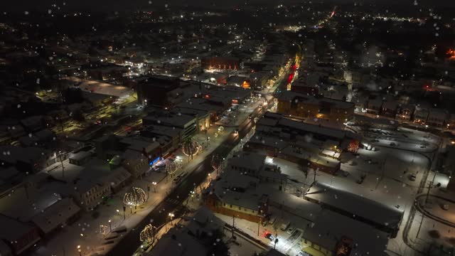 American town main street decorated with festive Christmas lights. Snow flurries falling on quaint town in America during winter night. High aerial orbit.