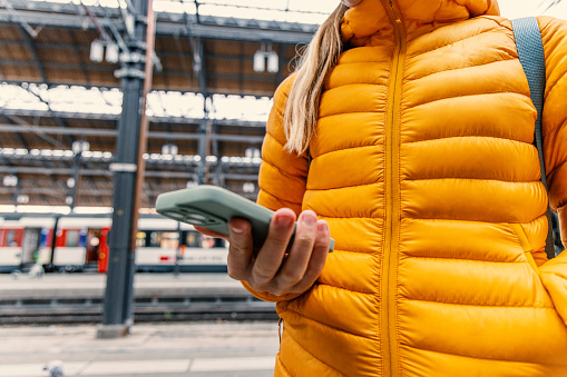 Close up shot of an unrecognized lady  with blonde hair in a yellow jacket typing on her mobile device with her right hand while keeping her left in the jacket's pocket.