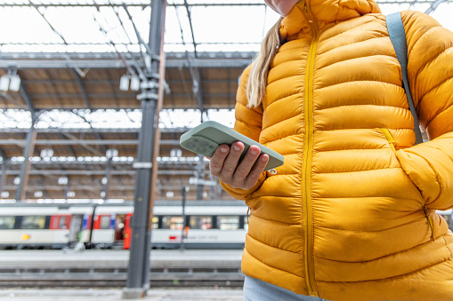 An unrecognized lady  with blonde hair in a yellow jacket holds her mobile device with her right hand while keeping her left in the jacket's pocket.
