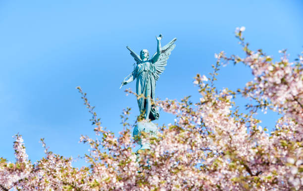Angelic statue for the fallen in war saluting high on a column above blooming pink cherry flowers commemorates the death of Admiral Ivar Huitfeldt (1886) in a Naval Battle or war. Copenhagen, Denmark