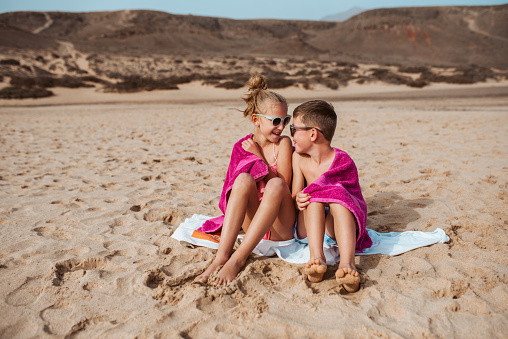 Siblings sitting on beach, wrapped in towel after swim in sea. Concept of family beach summer vacation with kids.