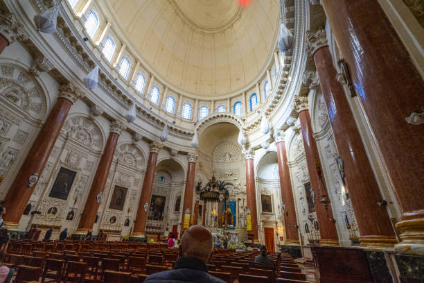 The Basilica of the National Shrine of the Blessed Virgin of Ta' Pinu is a Roman Catholic minor basilica and national shrine located near Garb on the island of Gozo, the sister island of Malta.\nIn 1883 a woman from the village of Garb, Karmni Grima, heard the voice of Our Lady at the small chapel that then occupied this site. It rapidly became a centre of pilgrimage and the number of visitors soon overwhelmed the little church.\nTodays magnificent church in Romanesque Style was built between 1920 and 1932. It is an architectural masterpiece, especially inside with its superb sculptures and craftsmanship in Maltese stone. The sanctuary was constructed in front of the original chapel. In 1932 the new Church was blessed and opened to the public and in 1935, Pope Pius XI raised it to the dignity of Minor Basilica.\nSince 2017 the space in front of basilica no longer serves as a carpark but with mosaic on both sides it became a place to welcome the devout apart from serving as an attraction and a spiritual aid to pilgrims. The mosaic work was commissioned to the Centro Aletti of Rome, under the guidance of Fr Marko Ivan Rupnik. The Centro Aletti is renowned for the sacred art which it has created in 150 churches all over the world, including the Vatican and sanctuaries such as Lourdes and Fatima.\nTa' Pinu, Gharb, Gozo, Malta, Europe.