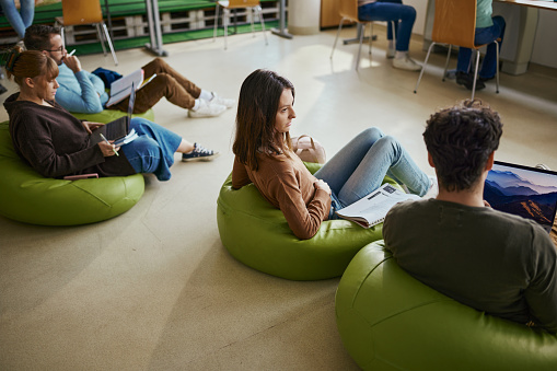 Young female student and her classmates relaxing on bean bags in campus. Copy space.