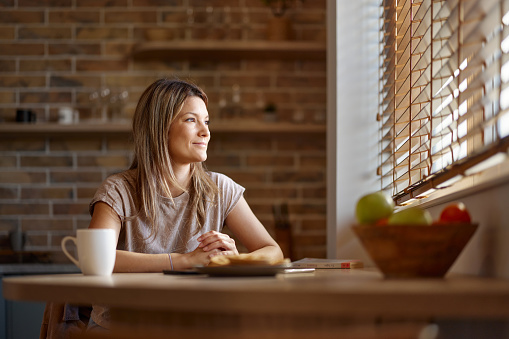 Smiling woman day dreaming while sitting at dining table and looking through window.