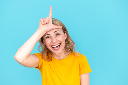 Smiling young woman making loser gesture over head. Playful female doing funny and comic face, fooling, enjoying goofy joke. Comic isolated over blue studio background with copy psace