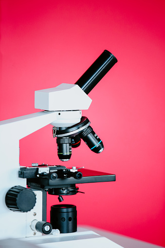 Close-up view of a microscope on a pink background. The microscope is a traditional design with a round base, a tall arm, and a binocular eyepiece