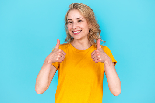 Happy woman in yellow t-shirt looking at camera, showing thumb-up gesture, approve deal, recommend offer, praises and supports good job, standing isolate on blue background with big smile