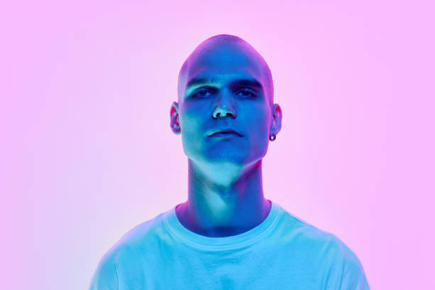 istock Portrait of serious young man with bald head, shaved face and ear piercing posing on gradient blue pink background in neon light 2148376103