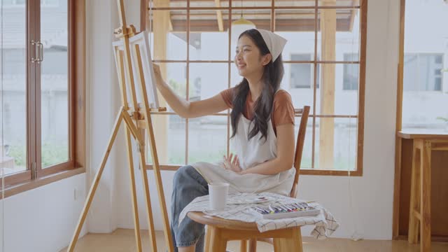 Asian young talented woman artist coloring on painting board in house. Attractive beautiful female draw art picture, creating artwork with watercolor paint and brush enjoy creativity activity at home