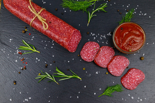 Salami sausage with ketchup and spices on black slate board. Top view