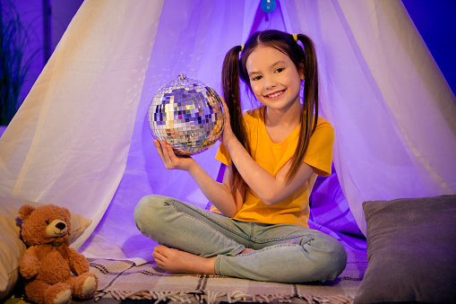 Photo portrait of cute small girl sit floor night light tent hold discoball dressed stylish yellow clothes modern playroom interior.