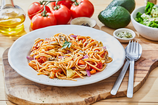 Healthy Vegan pasta, made of zucchini, mushrooms, Spanish onion and garlic, served on an elegant ceramic plate, with fresh vegetables and herbs around, avocado, cherry tomato, olive oil and pepper, on a light wooden natural kitchen or restaurant table, representing a wellbeing and a healthy lifestyle and food indulgence, top view image with a copy space