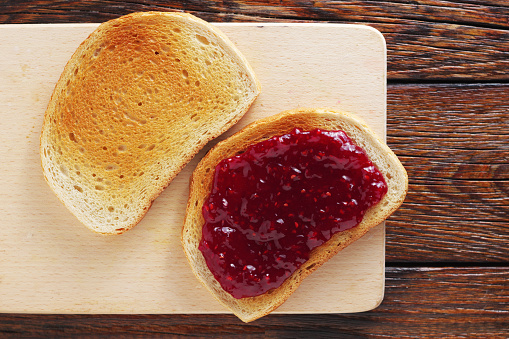 Two slices of toasted white bread, one with jam, the other without, on a cutting board, on old wooden background, top view
