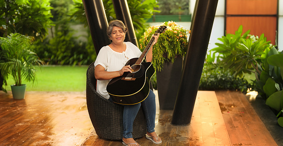 Indian Hindu happy mature middle aged older woman sitting on chair singing song play acoustic guitar enjoy music spend time outdoor home. Beautiful smiling singer old elder female guitarist practice tuning on musical instrument outside park