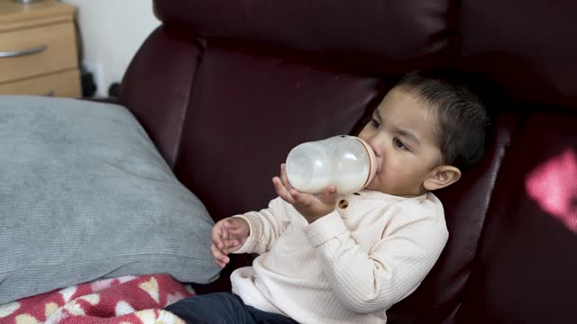 18-month-old toddler drinking milk from bottle sitting on sofa, capturing a timeless concept of nurturing and nourishment. Slow motion shot
