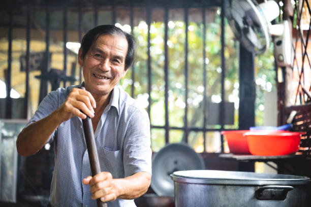senior man stirring in hot caramel for candy production on open fire in vietnam, looking at camera