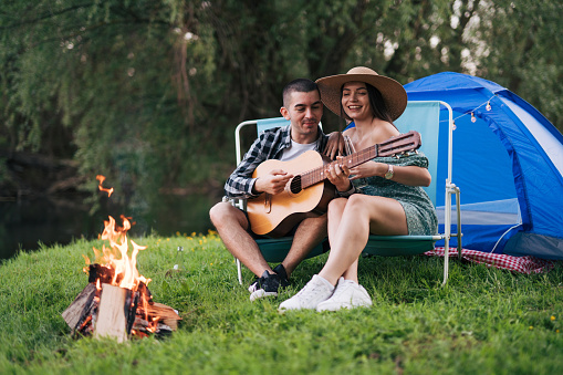 Young man playing the guitar to his girlfriend while they are camping. They enjoy nature and sit by the fire.
