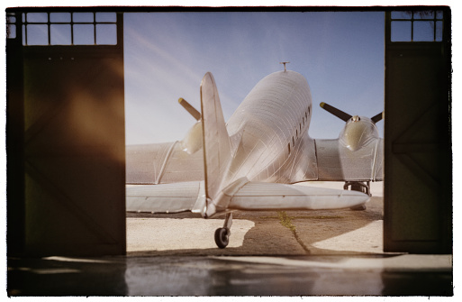 An iconic bare-metal DC-3 Dakota aeroplane sits outside of an old-style aircraft hangar. Model photography.