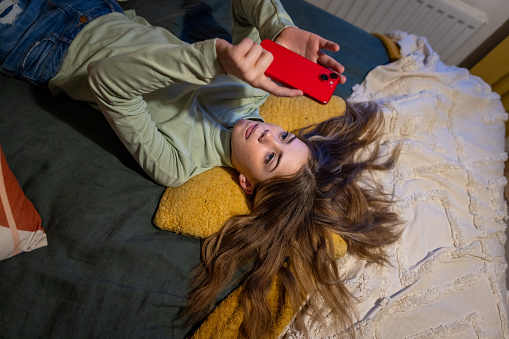 A high-angle mid-shot of a young girl lying on top of a bed on her back, she is concentrating and holding her phone above her head browsing social media. The location is their home located in North East of England in Newcastle Upon Tyne.