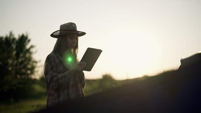 Farm worker woman with tablet in hands walks on agricultural grow field early spring at sunset. Blonde female works on farmland outdoors. Food production, farming industry, agribusiness concept.