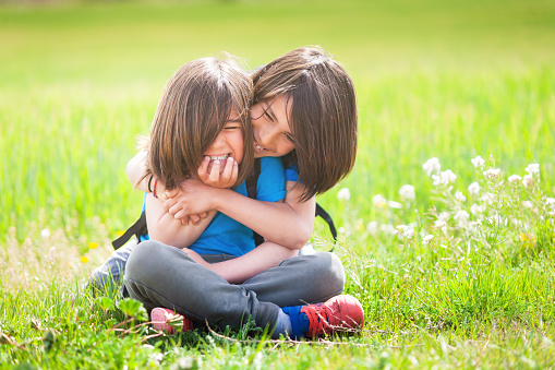 Big brother affectionately hugs his little brother in the middle of a green meadow with flowers on a sunny spring day. On his face there are big smiles and they reflect happiness and love.