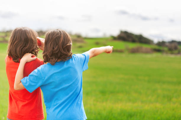 Two twin brothers with their backs to the camera, looking at the spring landscape of green meadows. One looks through binoculars and the other points to something on the horizon.
