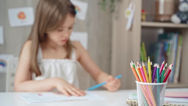 A close-up of colored pencils on the desk, with a creative young girl drawing in the background