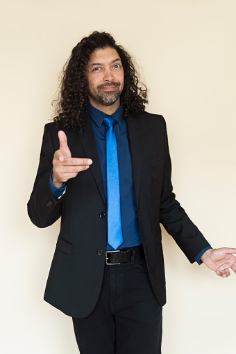 Portrait of stylish modern professional multiracial man on beige background. He is in his forties, wearing a blue shirt, a light blue tie and black suit. He has long curly hair and a beard. He is standing and looking at the camera. Vertical three quarter length studio shot with copy space. This was taken in Montreal, Quebec, Canada.