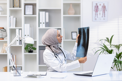 Focused muslim woman doctor with stethoscope checking x-ray of lungs while sitting at hospital cabinet with laptop. Qualified pulmonologist in white medical uniform making diagnosis for patient.